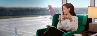 Eine stilvoll gekleidete Frau sitzt mit einer Tasse Kaffee in den Händen in einem grünen Sessel an einer Glasfassade in einer Flughafen Lounge und blickt entspannt hinaus aufs Rollfeld unter blauem Himmel.