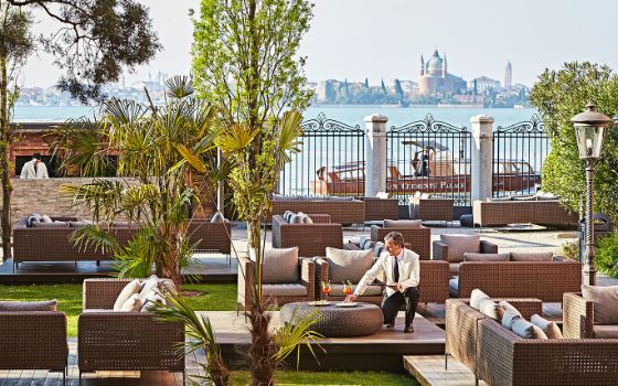 Ein Kellner stellt Cocktails auf einen Tisch auf einer luxuriösen Terrasse mit zahlreichen Sitzgelegenheiten und Bootsanleger an einer Lagune mit Blick auf Venedig. 