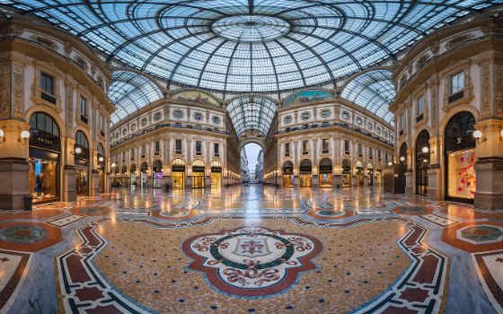 Weitwinkelaufnahme einer eleganten, menschenleeren Einkaufspassage mit Mosaikboden unter einem Glaskuppeldach am Abend. 