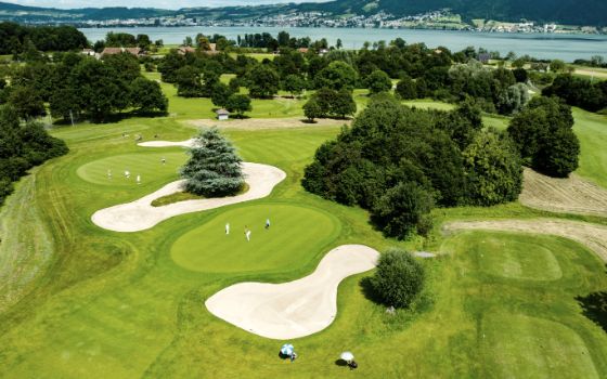 Luftaufnahme verschiedener Golfbahnen des Golfparks Holzhäusern mit Fairways, Grüns, aktiven Spielerinnen und Spielern und See im Hintergrund.
