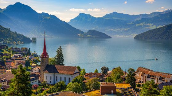 Aussicht auf den Vierwaldstättersee mit Kirche und idyllischem Dorf im Vordergrund, umgeben von den Schweizer Alpen, exklusives Reiseziel mit American Express Vorteilen.