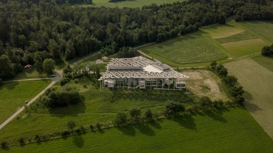 Luftaufnahme eines grossen, weissen modernen Gebäudes mit organischer Deckenkonstruktion inmitten von Feldern und Bäumen.