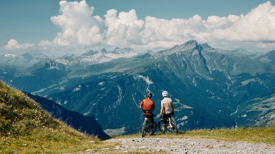 Zwei Männer auf Mountainbikes stehen auf einer Bergstrasse und schauen auf eine Berglandschaft.