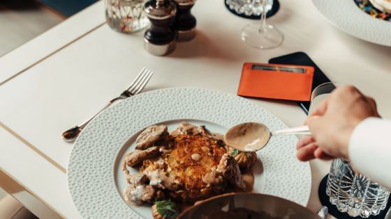 Eine Hand richtet mit einem Löffel ein Gerichtet auf einem weissen Teller auf einem edel gedeckten Restauranttisch an, daneben liegt eine silberne Kreditkarte von American Express in einem Etui.