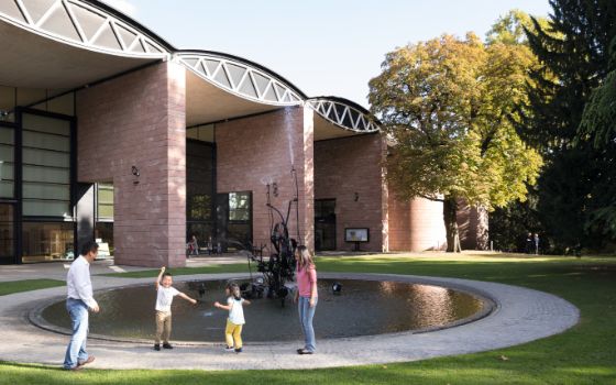 Zwei Erwachsene mit zwei fröhlichen Kindern vor dem Eingang eines Museums an einem runden Brunnen, der von einer Wiese umgeben ist. 