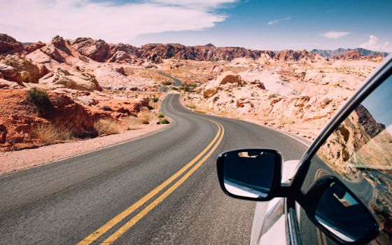 Auto mit Aussenspiegel im Anschnitt auf einer Strasse mit gelber Markierung in einer Wüstenlandschaft mit rötlichen Felsen