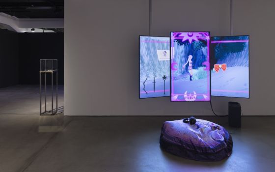 Abgedunkelter Raum in einem Museum mit dezenter Beleuchtung. An einer Wand sind mehrere Bildschirme angebracht, die eine bunte Grafik anzeigen, davor ein Sitzsack auf dem Boden, auf dem Kopfhörer und ein Controller liegen.