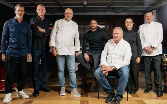Das Porträt zeigt sieben Küchenchefs in Kochjacken, die teilweise auf Stühlen sitzen und teilweise stehen.