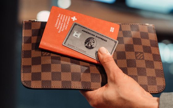 Eine silberne Kreditkarte von American Express und ein roter Schweizer Reisepass auf einem Lederetui, hochgehalten von einer Hand