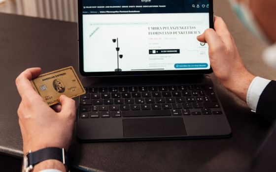 Zwei männliche Hände halten eine goldene American Express Kreditkarte und einen Laptop, auf dem Bildschirm ein Online-Shop.