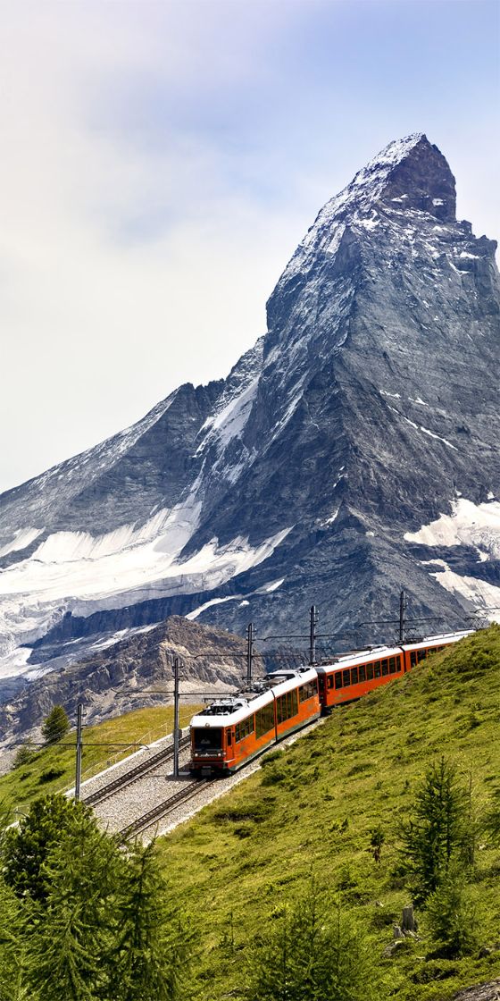 Eine rote Bergbahn fährt auf einer Trasse über einen grasbewachsenen Bergkamm vor der Kulisse eines schneebedeckten Berggipfels.