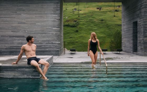 Ein Mann und eine Frau am Einstieg eines modernen Aussenpools, gerahmt von einer Betonfassade mit Durchblick auf eine Grasfläche am Berghang.