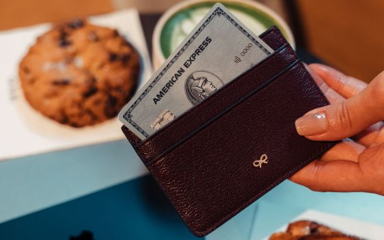 Nahaufnahme einer silbernen Kreditkarte von American Express in einem braunen Lederetui, gehalten von einer Hand, über Backwaren und einem Matcha-Getränk auf einem Tisch.