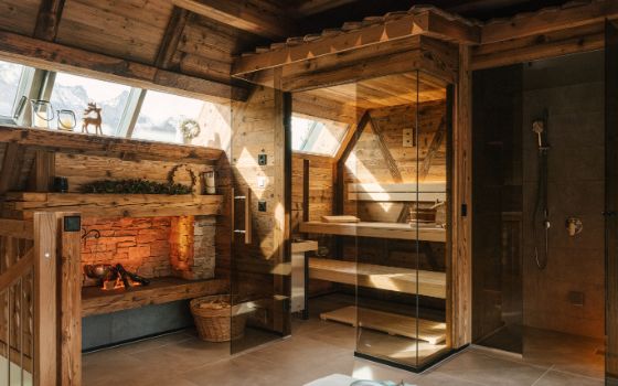 Ein mit Holz verkleideter Raum mit einer kleinen Sauna mit Glastüre, daneben ein Kamin mit Feuer.