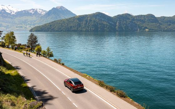 Auf einer Strasse in den Bergen fährt ein rotes Auto an einem grossen See entlang, im Hintergrund ragt eine Berglandschaft empor. 