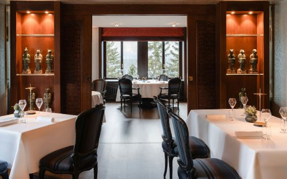 Heller, eleganter Speisesaal mit weiss eingedeckten Tischen und Skulpturen in beleuchteten Holzvitrinen, im Hintergrund ein Panoramafenster mit Blick auf Waldlandschaft.