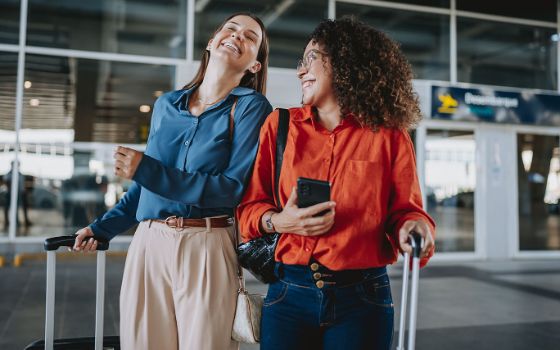 Zwei gut gelaunte Frauen stehen mit Koffern vor dem Eingang eines Flughafenterminals.
