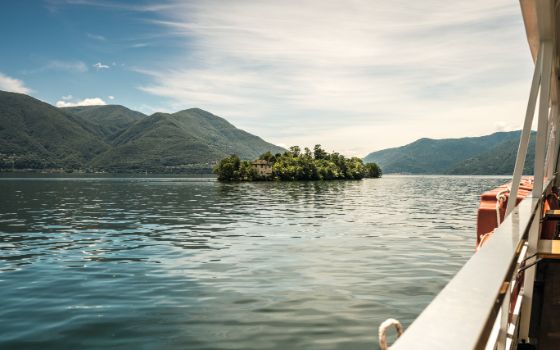 Blick von einem Boot auf einen See mit einer kleinen, grünen Insel, im Hintergrund grün bewachsene Berge. 