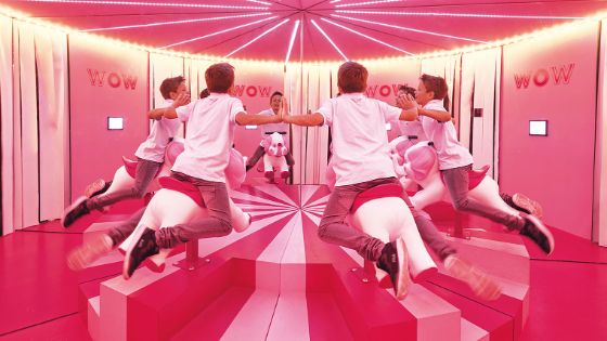 Ein Junge sitzt auf einer Elefantenfigur auf einem Trapez mit ausgestreckten Armen in einem Raum mit Spiegeln, der eine perspektivische Illusion erzeugt.