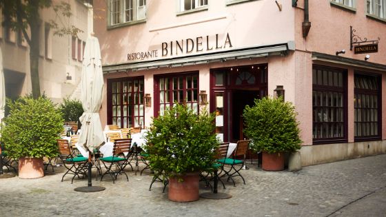 Aussenfassade des Restaurants Bindella mit kleiner, bestuhlter Terrasse auf Kopfsteinpflaster in Innenstadtlage.