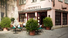 Aussenfassade des Restaurants Bindella mit kleiner, bestuhlter Terrasse auf Kopfsteinpflaster in Innenstadtlage.