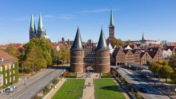 Personen in einer Parkanlage vor einem historischen Stadttor im spätgotischen Stil mit Doppelturm samt Kegeldächern vor der Altstadt in Lübeck bei Sonnenschein. 
