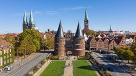 Personen in einer Parkanlage vor einem historischen Stadttor im spätgotischen Stil mit Doppelturm samt Kegeldächern vor der Altstadt in Lübeck bei Sonnenschein. 