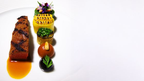 Auf einem weissen Teller liegt ein gebratenes Stück Fleisch mit brauner Sosse, daneben sind verschiedene Gemüse drapiert und mit Kräutern und Blumen verziert.