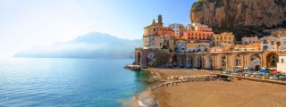 Mediterraner Fischerort an einer Felsküste mit bunten Häusern und Bogenbrücke entlang einer Bucht mit Sandstrand.