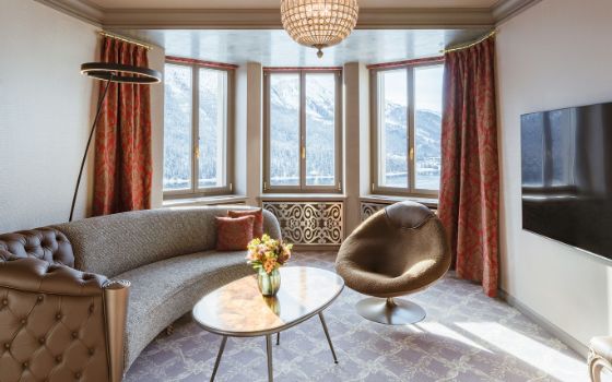 Helles, elegantes Wohnzimmer mit gerundeter Couch und Flachbildfernseher vor einer Fensterfront mit Blick auf einen See vor verschneiter Berglandschaft.
