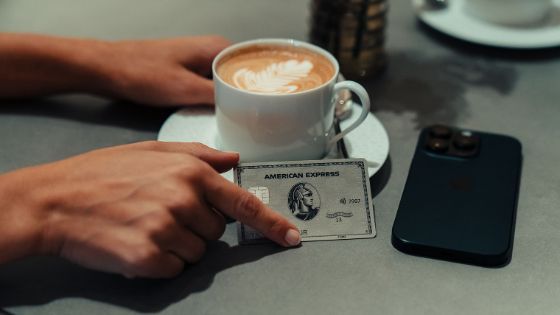 Zwei Hände umfassen eine weisse Cappuccino-Tasse und eine silberne Platinum Kreditkarte von American Express auf einem Tisch, daneben liegt ein Smartphone. 