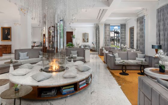 Helle, moderne Hotellobby mit zahlreichen Sitzgelegenheiten in Grautönen und einem freistehenden, gläsernen Kamin. 