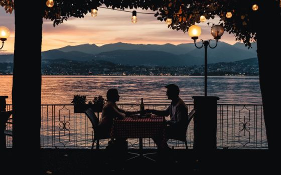 Silhouette zweier Personen an einem Tisch auf einer Terrasse unter Bäumen mit Lichterkette an einem See bei Sonnenuntergang. 