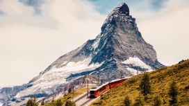 Eine rote Bergbahn fährt auf einer Trasse über einen grasbewachsenen Bergkamm vor der Kulisse eines schneebedeckten Berggipfels.