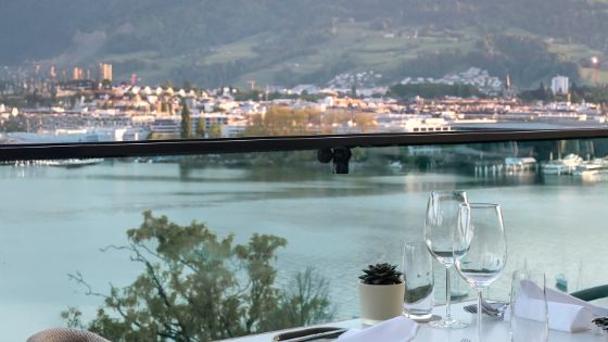 Gedeckter Restauranttisch mit Weingläsern, dahinter die Aussicht auf den Vierwaldstättersee und verschwommen die Stadt Luzern. 