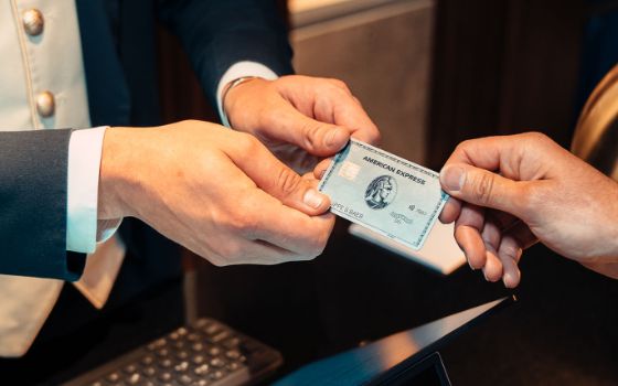 Eine silberne Kreditkarte von American Express wird von einer Hand in zwei Hände einer Person in eleganter Uniform an einer Rezeption übergeben. 