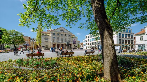 Belebter Platz mit Blumenbeeten und Goethe-Schiller-Denkmal vor einem Theatergebäude im Stadtzentrum von Weimar.