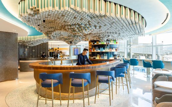 Mitarbeiter hinter einer runden Bartheke unter einer modernen, skulpturalen Deckenkonstruktion in einer hellen Flughafen-Lounge.