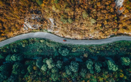 Sicht auf ein rotes Auto aus der Vogelperspektive, es fährt auf einer Straße durch ein waldreiches Gebiet.