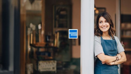 Eine junge Frau in dunkler Schürze steht mit verschränkten Armen lächelnd an einem Rahmen vor einer Fensterfront, an einem Fenster klebt das Logo von American Express.