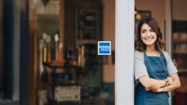 Eine junge Frau in dunkler Schürze steht mit verschränkten Armen lächelnd an einem Rahmen vor einer Fensterfront, an einem Fenster klebt das Logo von American Express.