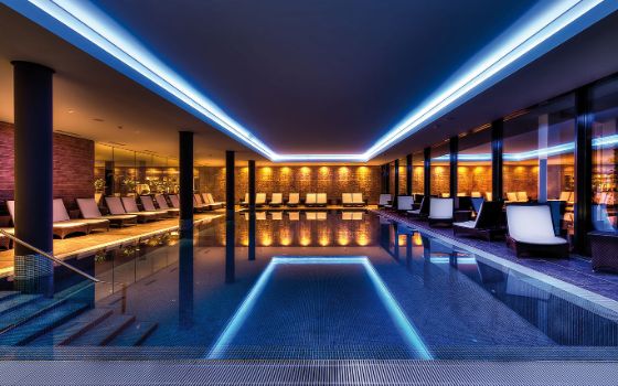 Ein grosser Innenpool ist blau beleuchtet, darum herum sind Liegen aufgestellt, die gelb angeleuchtet werden. 