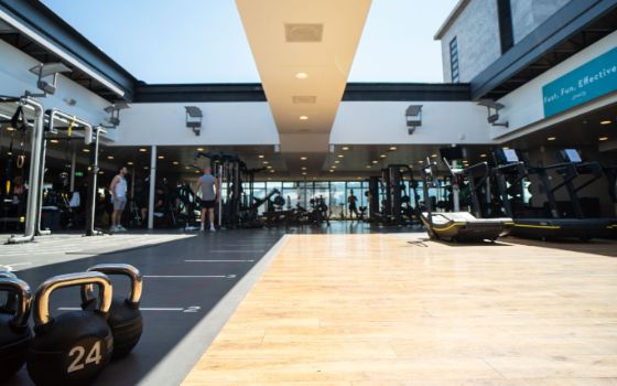 Personen in einem Fitnessstudio mit einer grossen Trainingsfläche unter einem geöffneten Dach bei Sonnenschein.