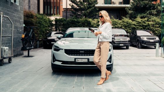 Frau in schicker Kleidung vor einem glänzenden Auto, in der Hand ein Handy, im Hintergrund weitere Autos und ein Kofferwagen.