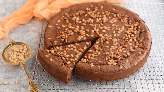 Ein angeschnittener runder, brauner Kuchen mit Pinienkernen auf einem Gitterrost. 