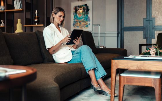 Eine moderne, stilvoll gekleidete Frau sitzt auf einem Sofa in einer Hotellobby und schaut auf ein Tablet in ihren Händen.