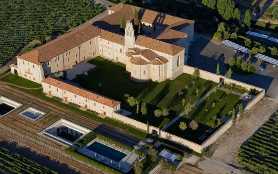 Luftaufnahme einer historischen Klosteranlage aus Naturstein, umgeben von großflächigen Rebflächen im Sonnenlicht. 