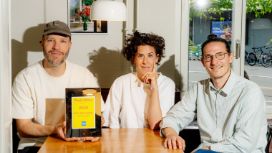 Drei Personen sitzen an einem Tisch im Restaurant Olympia in Bern. Die Person links hält eine GaultMillau-Auszeichnung mit dem Titel „POP DES JAHRES 2026“ in den Händen. Alle drei blicken lächelnd in die Kamera. Im Hintergrund sind ein Fenster, Pflanzen und ein Strassenschild zu sehen.