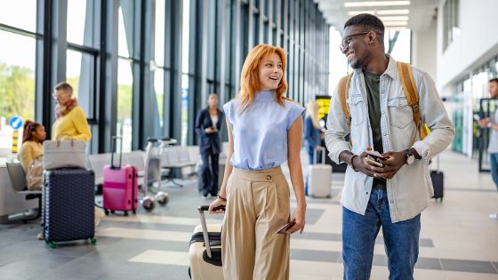 Zwei Personen laufen mit Rollkoffer und Rucksack in einem Flughafen entlang. 