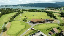 Anlage des Golfparks Holzhäusern von oben mit Fairways, Grüns und Driving Range, im Hintergrund ein See und Berge.
